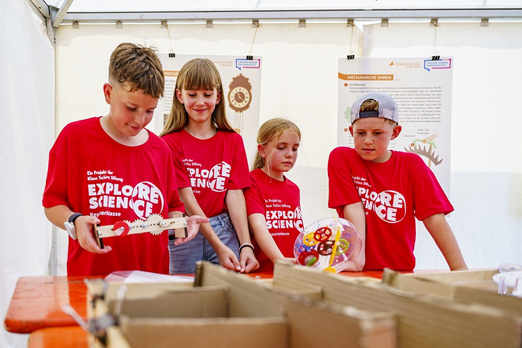 Vier Kinder in roten Explore Science-T-Shirts präsentieren bei einem Wettbewerb selbst gebaute Konstruktionen auf einem Tisch. Im Hintergrund sind Informationstafeln und ein Veranstaltungszelt zu sehen.