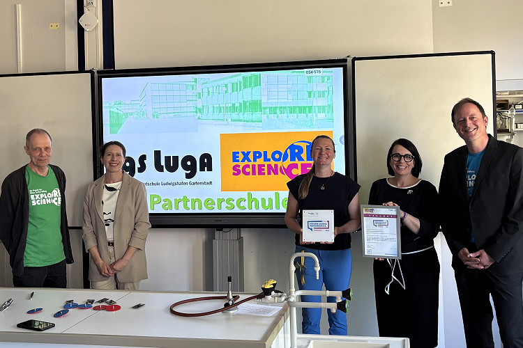 Fünf Personen stehen vor einem Bildschirm mit dem Explore Science-Logo und der Aufschrift „Partnerschule“ in einem Klassenraum. Zwei Frauen in der Mitte halten Urkunde und Plakette zur Auszeichnung als Explore Science-Partnerschule. Auf dem Tisch im Vordergrund liegen Unterrichtsmaterialien. Von links nach rechts: Lehrer Gerd Hegeler-Burkhart, Lehrerin Anne Witzel, Désirée Pschorn, Lilian Knobel (Klaus Tschira Stiftung), Schulleiter Thomas Lück.